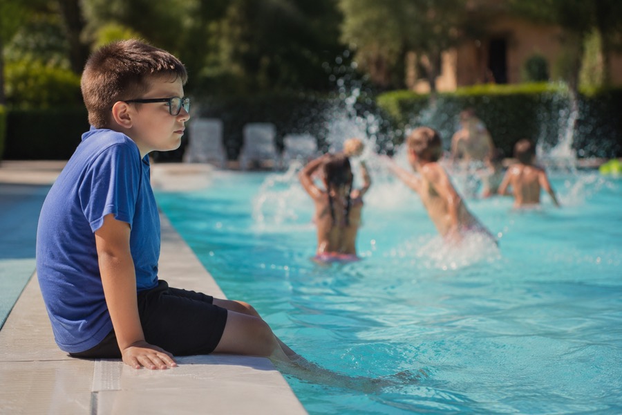 Copil cu ochelari stând pe marginea piscinei, în timp ce ceilalți copii se joacă în apă